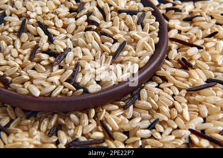 A mixture of brown and black unpolished wild rice in a clay ceramic bowl close-up against the background of scattered grains of rice Stock Photo