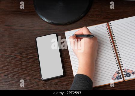 female hands, a girl writes in a notebook, uses a black smartphone, desktop, dark background with copy space, for advertising, top view, white box nic Stock Photo
