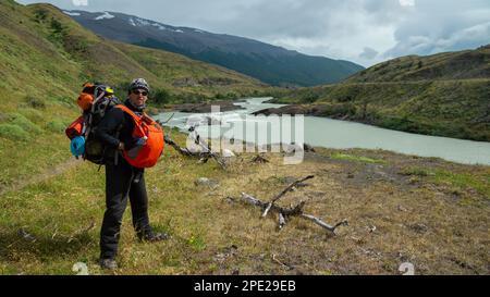 Young tourist carrying two backpacks posing facing forward next to a river with green mountains in the background on a cloudy day Stock Photo