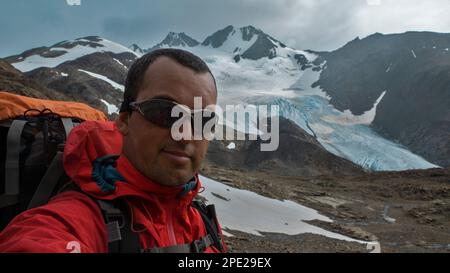 Young climber with red jacket and big backpack taking a selfie against mountains with glaciers in the background during a cloudy day Stock Photo