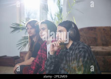 Three men with mobile phones and headphones enjoy music at home. A group of three young people feeling free, enjoying life. Stock Photo