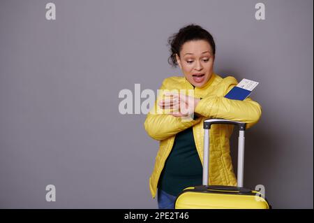 Multi-ethnic woman in yellow jacket, checks time on her wrist watch, feeling anxious being unpunctual at her flight Stock Photo