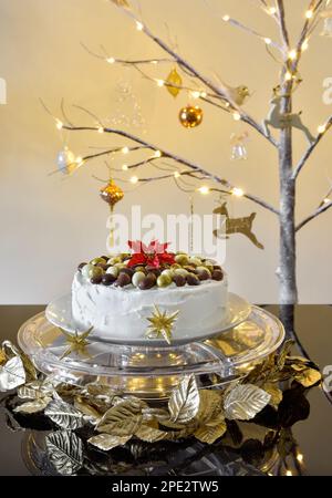 Iced Christmas cake with chocolate nuts on top on clear cake stand with a lit twig Christmas tree in the background Stock Photo
