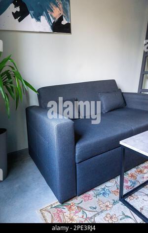 A small modern blue and gray sofa, next to a palm tree in a tub and a small loft-style table with a marble top Stock Photo