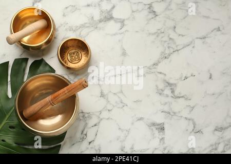 Golden singing bowls, mallets and monstera leaf on white marble table, flat lay. Space for text Stock Photo