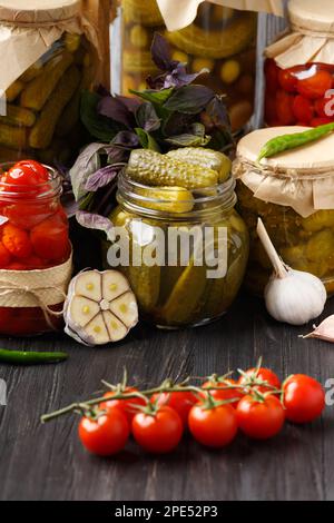 Pickled cucumbers and tomatoes in closed and open jars, fresh cherry tomatoes on a branch, garlic, chili peppers, basil on a dark wooden background. Stock Photo