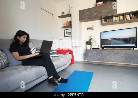 Young woman on a couch using a black laptop in her living room. Browsing internet. Woman working with the TV on. Work from home concept. Stock Photo