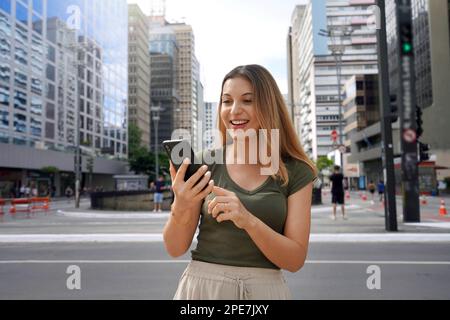 Cheerful Brazilian girl watching funny video on phone standing on street at daytime. Brown-haired with toothy smile wears green t-shirt. Social media Stock Photo