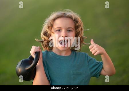 Child lifting the kettlebell in park outside. Sport activities at leisure with children. Sporty kid boy holding dumbbells kettlebell. Stock Photo