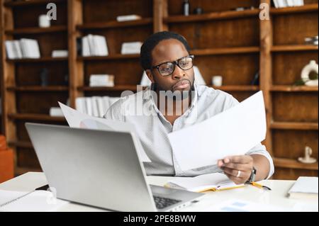 Clever and concentrated African-American guy is doing paperwork, a man looking through workpaper sitting at the desk with a laptop in office, a man checking report, collate data Stock Photo