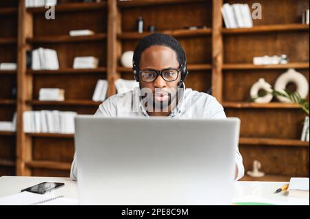Serious african-american businessman in eyeglasses sitting in front of the laptop. Puzzled male employee staring at the screen, thinking of difficult decision, solving task at office. Stock Photo
