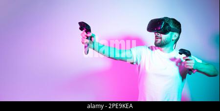 Bearded man in VR glasses plays in simulation figth and using controllers in gradient neon background. Concept interactive modern online game Stock Photo