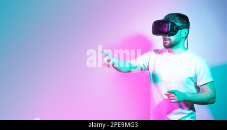 Bearded man in vr glasses plays and tries to touch something in gradient neon background. Augmented reality technology,online game, entertainment Stock Photo