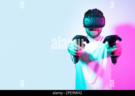 Bearded, smiling middle age man in VR glasses using controllers in multicolored pastel neon colorful background. Interactive innovation online game Stock Photo