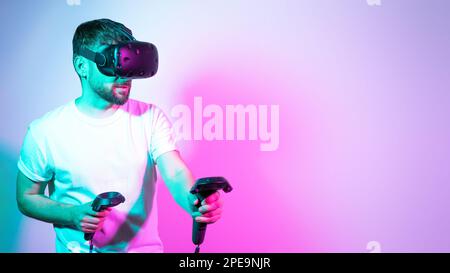 Concept modern hobby. Bearded millennial man in VR glasses plays in simulation figth and using controllers in gradient neon background. interactive Stock Photo