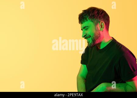 Creative portrait. Smiling man with closed eyes in green, pink neon lights on yellow background. Modern photo handsome bearded middle age man Stock Photo