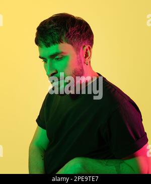 Artistic creative portrait. Pensive handsome man looking down in green, pink neon lights on yellow background. Modern photo of bearded middle age man Stock Photo