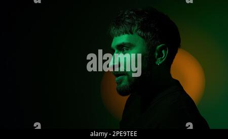 Side view artistic creative portrait of man looking away in orange neon lights on dark green background. Modern photo handsome bearded middle age man Stock Photo