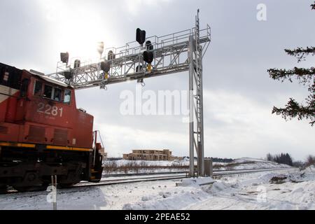 A red locomotive on passes under railway signal lights as the sun shines through clouds on a winter day, snow on the ground. Stock Photo