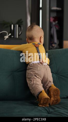 Back view of an infant kid in yellow clothes standing on green sofa Stock Photo