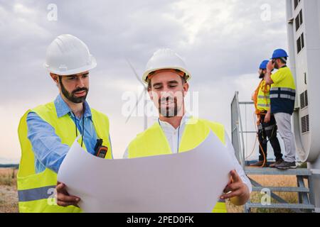 Two windmill engineers looking a blueprint with the plan of the construction of the electricy turbine. At background workers or contractors doing their job . Renewable energy concept. High quality photo Stock Photo