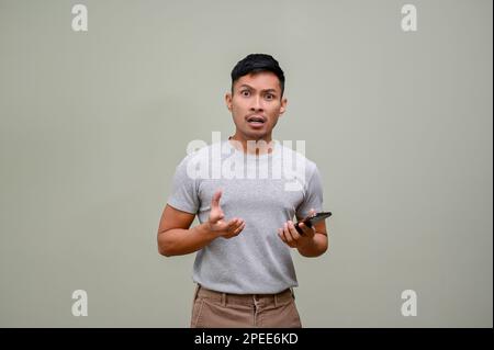 Shocked millennial Asian man in casual clothes standing against an isolated green background with his phone in his hand. Stock Photo