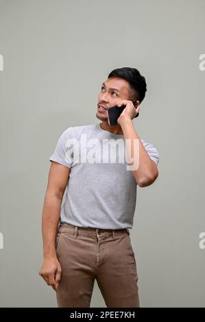 Confused millennial Asian man in casual clothes looking aside or looking up and talking on the phone with someone while standing against an isolated g Stock Photo