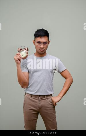 Serious and stressed millennial Asian man in casual clothes and glasses holding an alarm clock while standing against a green studio background. on ti Stock Photo