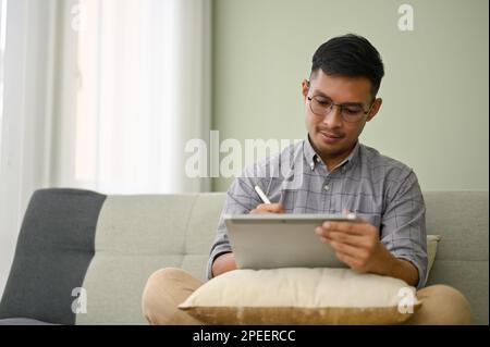 Handsome and focused millennial Asian man in casual clothes using his digital tablet on sofa in his living room. Stock Photo