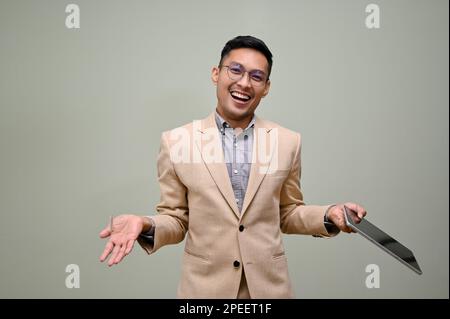 Cheerful and smiling millennial Asian businessman in a formal business suit holds a tablet, opens his palms, and looks at the camera while standing ag Stock Photo