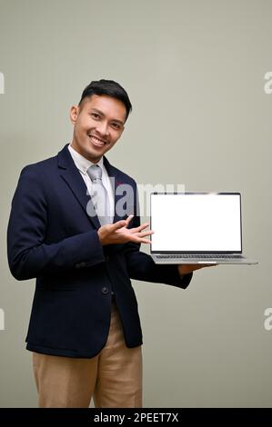 Handsome adult Asian businessman in formal business suit, holding laptop white screen mockup, open his palm at the screen, standing against a green ba Stock Photo