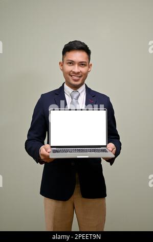 Handsome millennial Asian businessman holding a laptop, showing laptop blank screen mockup to camera while standing against an isolated green backgrou Stock Photo