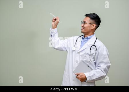 Smart and cheerful millennial Asian male doctor in white gown with his portable tablet and stethoscope is pointing a pen at the empty space, isolated Stock Photo