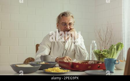 Elderly retired grandfather stay at home with unhappy face sitting alone on eating table in house. Depressed mature Senior old man upset feeling lonel Stock Photo