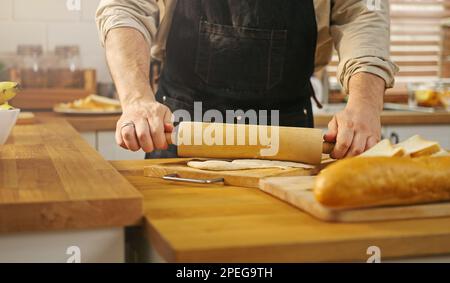 Close-up hands man wearing apron holding a rolling pin, making dough on kitchen table. Stock Photo