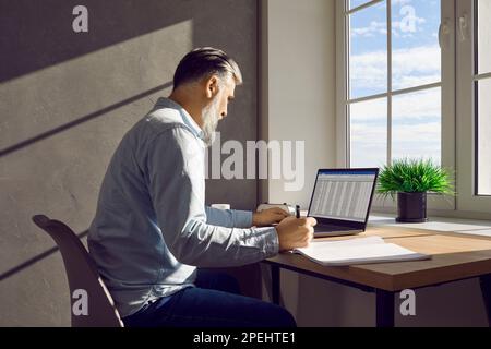 Side view of mature business man working on a laptop with tables at office and writing. Stock Photo