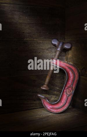 A rusty, red, vintage three inch steel C-Clamp on a rustic wooden setting in soft, dark mood lighting; captured in a studio Stock Photo