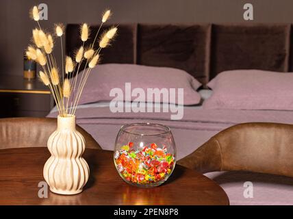 A figured earthenware vase with fluffy grass stalks and a glass vase with multi-colored sweets in a background blurred bedroom. Stock Photo