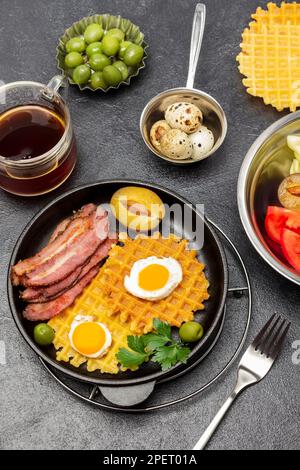 Waffles, fried bacon and eggs in a frying pan. A cup of tea, green olives and quail eggs in bowls. Flat lay. Black background Stock Photo