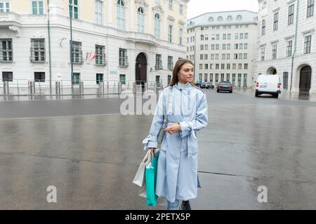 young woman in blue trench coat walking with shopping bags while walking near buildings in Vienna Stock Photo