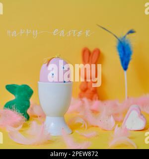 text happy easter and a pink easter egg in a white eggcup, some pink feathers and some easter bunnies of different colors on a yellow background Stock Photo