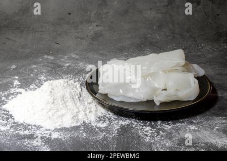 Front view of flat white big noodles uncooked in a black plate surrounded by all-purpose flour. on a dark background Stock Photo