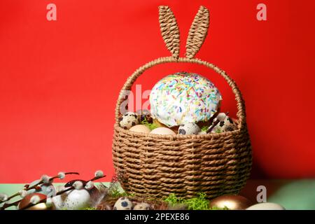 Easter celebration. Easter decorations in a basket with rabbit ears. Easter cake and eggs on moss, quail eggs and feathers on a red background. Tradit Stock Photo