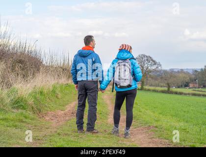 Rear view of a married, UK couple holding hands, dressed in warm hiking gear, looking over countryside fields into the distance. Stock Photo