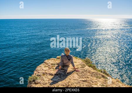 Back of young woman sitting on the top of cliffs immersed in the beauty of the Atlantic Ocean at Marinha Beach in Algarve, Portugal Stock Photo
