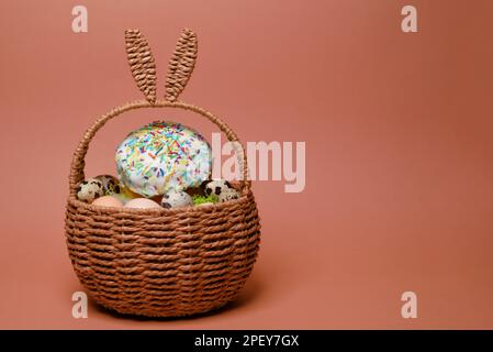 Easter decorations in a basket with rabbit ears. Easter cake and eggs on moss, quail eggs and feathers on a pink background. Place for text Stock Photo