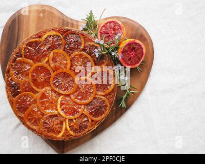 Blood orange upside-down cake and rosemary garnish shot from above on a heart-shaped serving board on a white background with space for copy Stock Photo