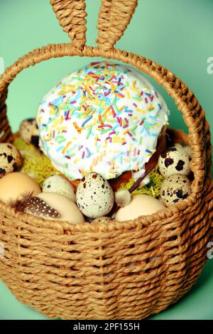 Easter decorations in a basket with rabbit ears. Easter cake and eggs on moss, quail eggs and feathers on mint green background Stock Photo