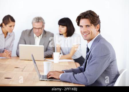 Confident, young male executive. Portrait of a handsome young executive working on his laptop with his team in the background. Stock Photo
