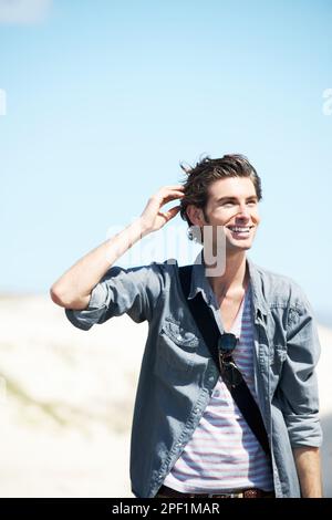 Travel broadens the mind. Handsome young man in a denim shirt smiling and touching his head with dunes and sky in the background. Stock Photo